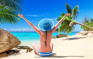 Vrouw in bikini met rug naar kijker toe zit op mooi zonnig strand, met palmbomen en rotsen. Ze kijkt richting de helderblauwe zee en heeft haar armen gespreid in de lucht. Ze is tevreden met de situatie