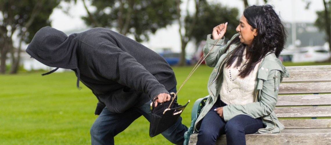 Niet stelen is het advies. De afbeelding laat een man in donkere kleding zien die de tas van een vrouw afpakt, terwijl zij op een bank in het park zit